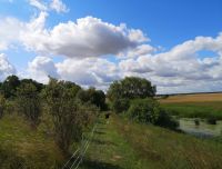 Skats uz Īslīces upes ieleju un saimniecības teritoriju, kurā pastaigājas saimniecības apmeklētājs un bauda lauku vidi.
